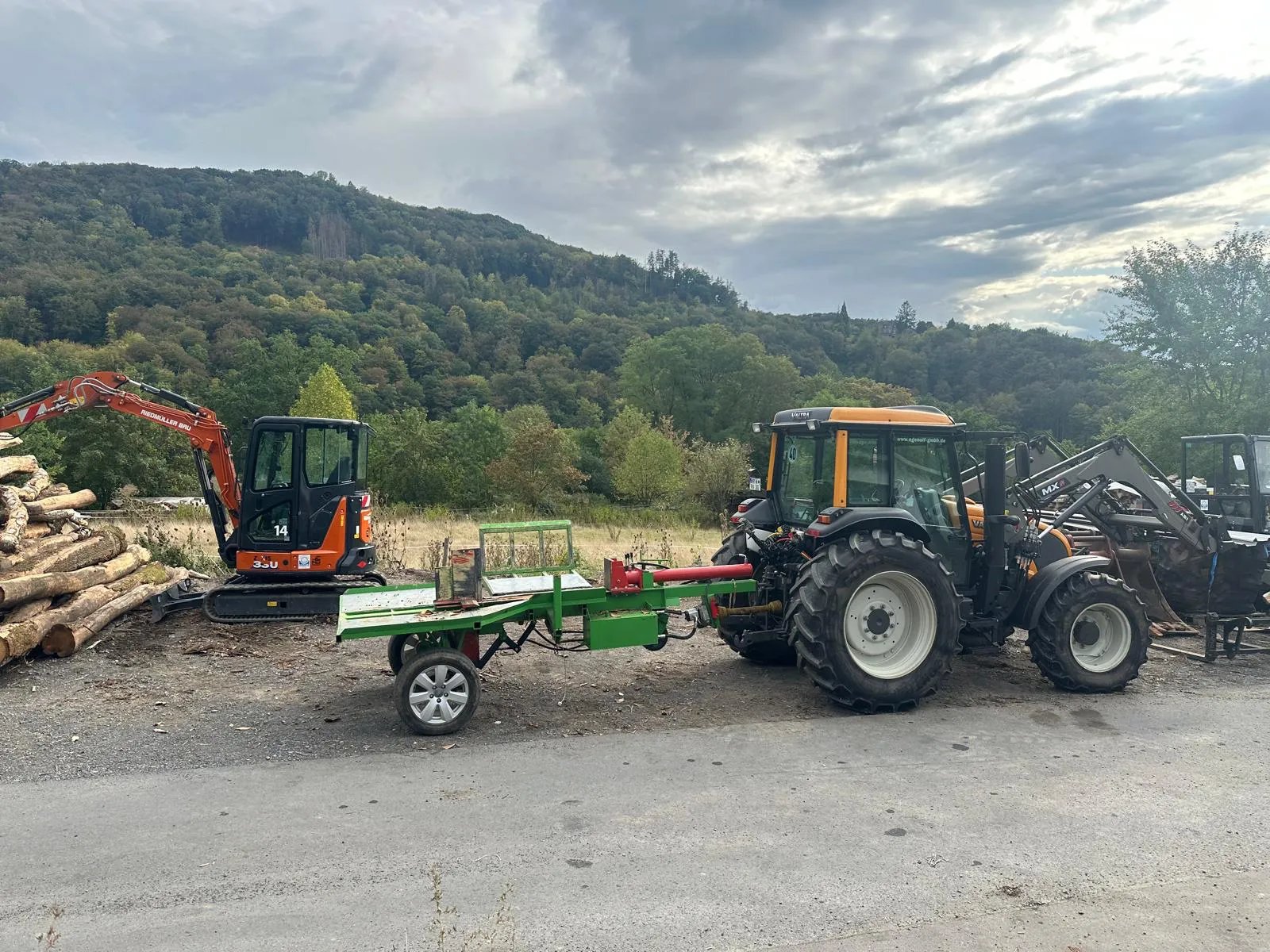 Traktor mit angehängtem Holzspalter auf dem Holzplatz, daneben ein Minibagger und Stammholz